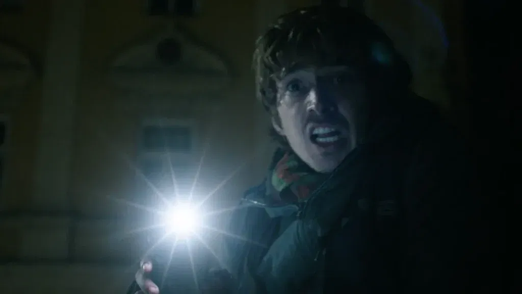 Resident Evil Ending Explained: What Does Bryan Become After Losing the Cure? 39 A wide-eyed man with a terrified, shouting expression holds up a flashlight that flares brightly in the darkness, wearing a scarf and jacket, with a dimly lit building visible behind him at night.