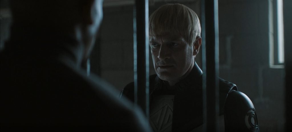 A blond man with a bowl haircut stares sternly through vertical prison bars. He wears a dark uniform with a Green Lantern emblem on the shoulder. The dark, out-of-focus back of another person's head fills the left foreground, and a bright window is faintly visible behind him.