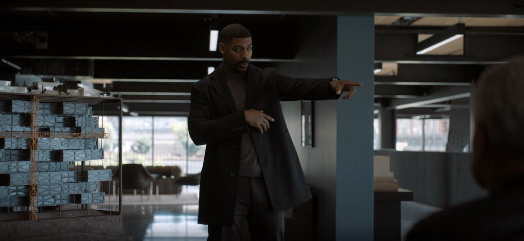 John Stewart, in a dark overcoat and grey turtleneck, points off-screen in a dim open-plan office. A blue-grey architectural building model stands at left.