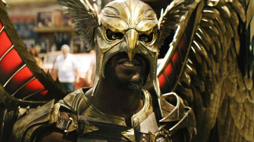 A figure in a weathered gold hawk helmet with a beak and feathered side wings grins at the camera, stubble visible below the mask. Large golden wings with red inner feathers spread out behind him, and he wears gold armor. A blurred street scene with a person in a white shirt is in the background.