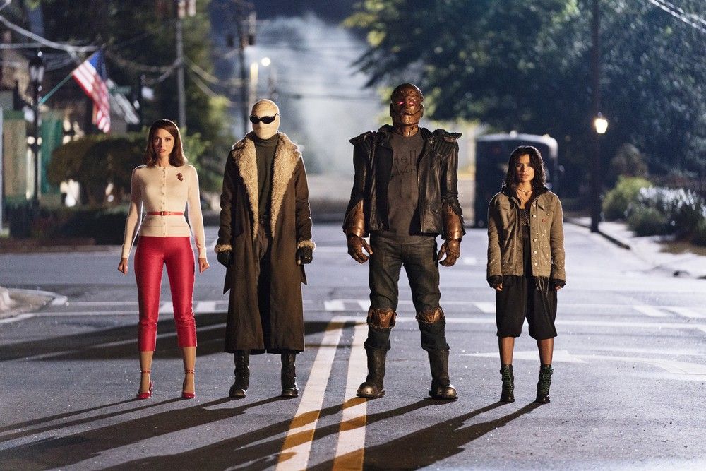 Four people stand side by side in the middle of a quiet small-town street at night. From left to right: a woman in a cream cardigan, red pants and red heels, a figure with a bandaged face, sunglasses and a long shearling-collared coat, a tall metallic figure with a glowing red eye in a leather jacket, and a woman with dark hair in a tan jacket, black shorts and green boots. An American flag and drifting mist are in the background.