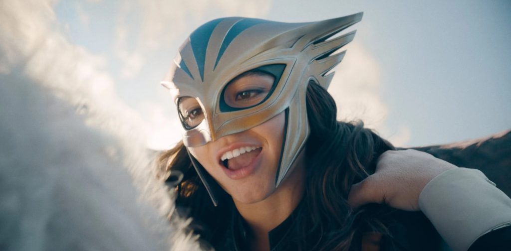 Low-angle close-up of a smiling woman with long dark hair, looking downward with her mouth open mid-laugh. She wears a silver and gold hawk-style mask with blue accents and swept-back wing details. A hand rests on her shoulder, and soft clouds fill the sky behind her.