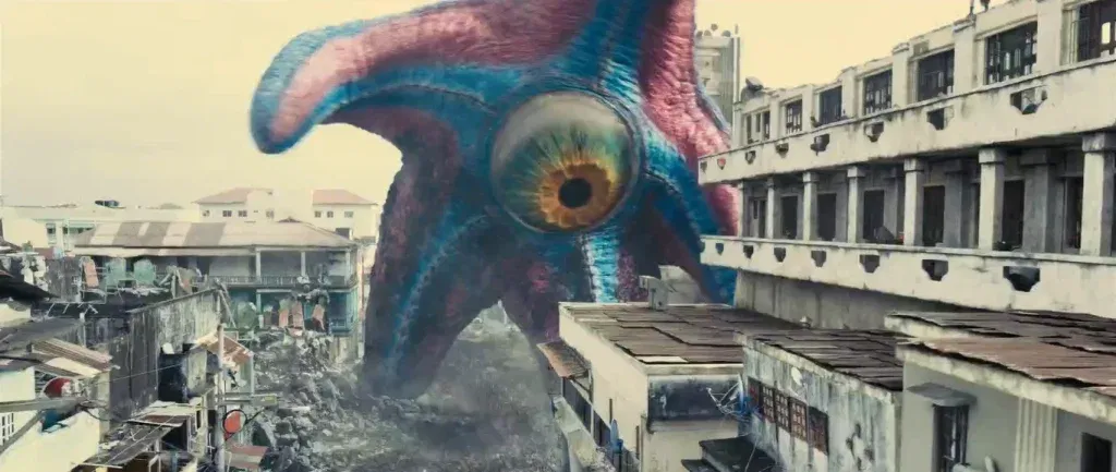 A colossal starfish-like creature with blue and pink skin and a single large eye with a green and amber iris looms over a crumbling city street. Its arm curls above weathered concrete buildings with corrugated metal rooftops, and rubble fills the road at its base. The sky is a hazy, pale yellow.