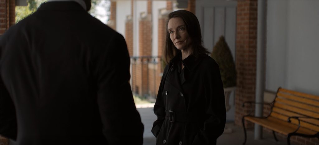 A woman with shoulder-length brown hair stands with her hands in the pockets of a black double-breasted trench coat, giving a faint, measured smile. She looks toward a man whose dark-suited back fills the left foreground. They stand under a covered brick walkway with white columns, and a wooden park bench and a potted topiary sit behind her.