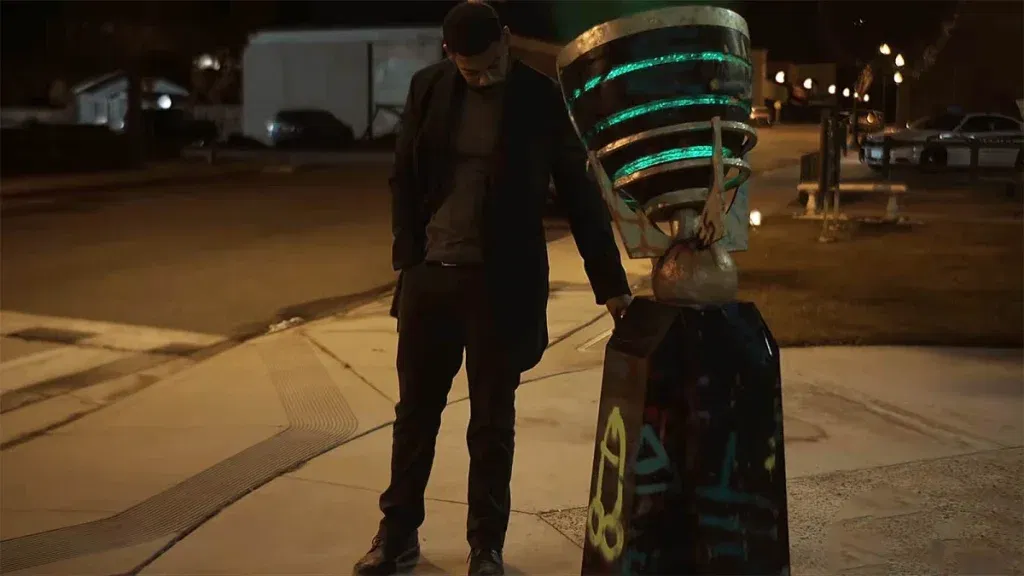 A man in a dark jacket, grey shirt and dark trousers stands on a nighttime sidewalk, head bowed, resting one hand on a tall stone monument. The monument is topped with a lantern-shaped sculpture with glowing green bands, and its base is covered in spray-painted graffiti, including a crude yellow doodle. A police car and a park bench sit in the background beneath distant streetlights.