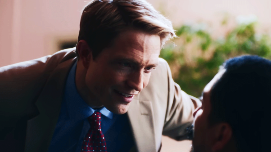 A man in a tan blazer, blue shirt, and red tie leans in close with a wide smile, hand on another person's shoulder, greeting them outdoors near green foliage. Robert Pattinson in primetime
