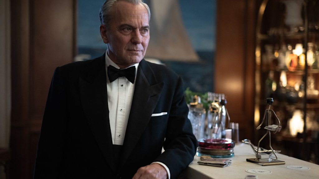 An older man in a black tuxedo and bow tie leans against a bar counter, gazing off to the side, with decanters and glassware visible in the background. A still from Netflix's The Final Problem