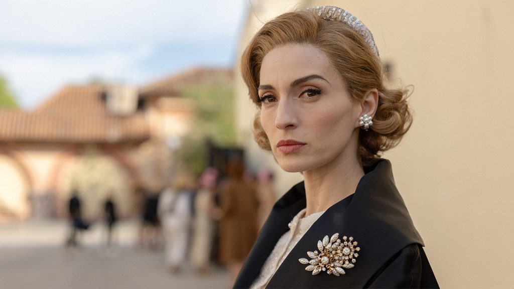 A woman with wavy auburn hair and a jeweled headband looks over her shoulder, wearing a black coat with a pearl brooch and pearl earrings, a sunlit town square blurred behind her.