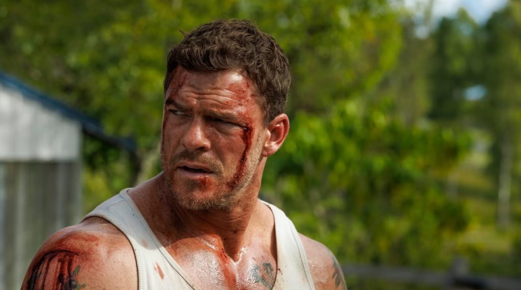 A bruised and bloodied man with curly hair and tattooed arms, wearing a white tank top, looks off to the side with a wary expression, standing outdoors in front of greenery.