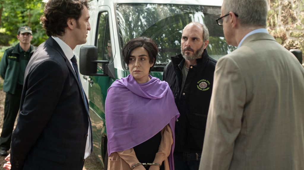 A handcuffed woman in a purple shawl stands beside a Guardia Civil police van in a wooded area, flanked by an officer in a Policía Judicial jacket and confronted by two men in suits, one in the foreground and one facing her. Netflix thriller series
