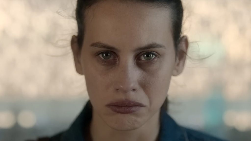 An extreme close-up of a woman's face with green eyes, dark circles, and hair pulled back, her expression haunted and unblinking against a hazy, sunlit background.