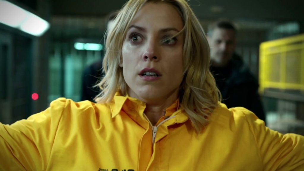 A blonde woman in a yellow prison jumpsuit stands with arms raised, her face tense and startled, as two guards look on behind her in a dimly lit corridor.