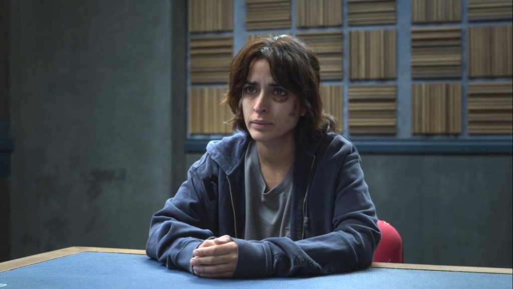 A young woman with disheveled hair and a bruised, tired face sits at a table in a soundproofed interrogation room, her hands folded in front of her as she looks up warily.