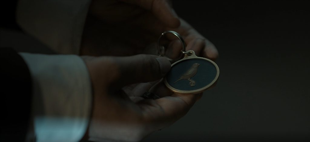 A close-up of a hand holding a round brass keychain fob with a small bird embossed on a dark blue enamel face, the metal ring looped over one finger, set against a shadowy, near-black background. A still from Lanterns episode 7