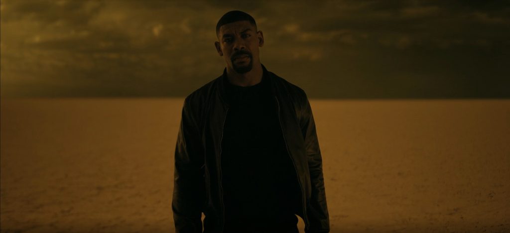 A man with a short fade and a goatee, wearing a black leather bomber jacket over a black tee, stands in near-silhouette on a vast, barren plain. The sky is a heavy, brooding mass of clouds, and the whole scene is bathed in a murky amber-gold haze. A still from lanterns
