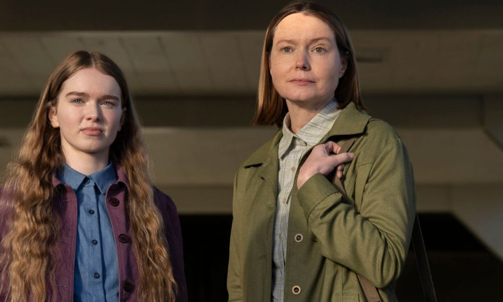On the left, a young girl with long, wavy auburn hair wears a purple jacket over a blue button-up shirt and looks ahead with a guarded expression. On the right, an older woman with shoulder-length auburn hair, in an olive green jacket over a plaid shirt, grips the strap of her bag and stares off with a stern, watchful look.
