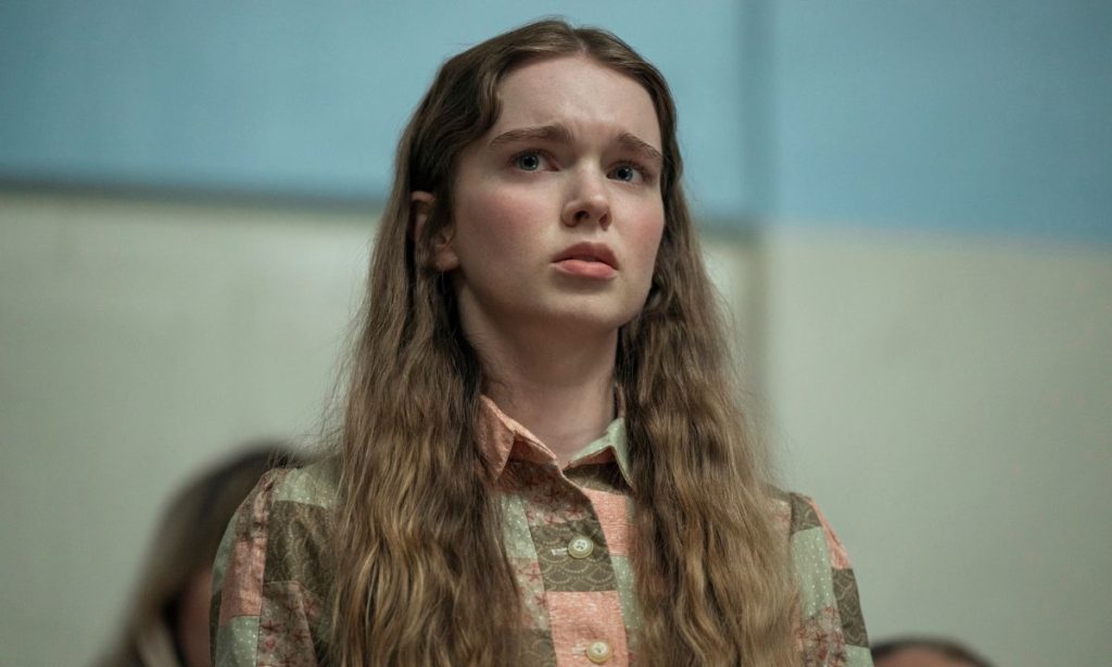 Carrie Review: All That Telekinesis, So Little Momentum 7 A teenage girl with long, wavy light-brown hair stands in a pale, tiled room, looking ahead with a worried, uncertain expression. She wears a patchwork-print collared shirt in muted green, peach, and brown, and other people are blurred in the background.
