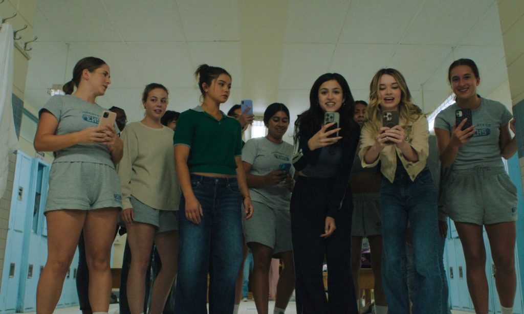 Carrie Review: All That Telekinesis, So Little Momentum 8 A group of teenage girls in a school locker room stand in a loose row, all pointing their phones toward the camera as if filming. Most wear gray gym clothes, and a few are in jeans and casual tops. Some smirk or look on with interest against a backdrop of pale blue lockers.