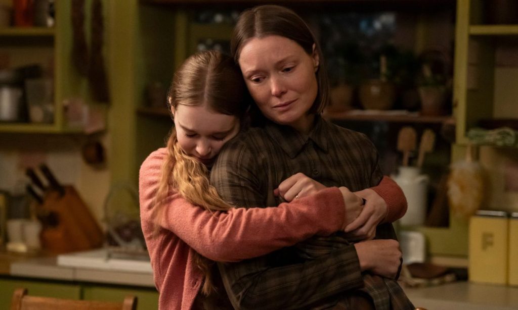 Carrie Review: All That Telekinesis, So Little Momentum 9 In a warmly lit, cluttered kitchen with olive-green shelves, a teenage girl in a coral cardigan hugs an older woman in a dark plaid shirt. The girl's eyes are closed and her face is pressed against the woman's shoulder, while the woman looks down with a pained, tender expression.