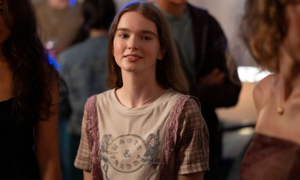 Carrie Review: All That Telekinesis, So Little Momentum 10 A teenage girl with long, straight light-brown hair stands in a dimly lit, crowded room, smiling faintly at the camera. She wears a cream graphic t-shirt with pink lace trim and plaid sleeves, and a thin necklace. Blurred partygoers fill the background. A still from carrie