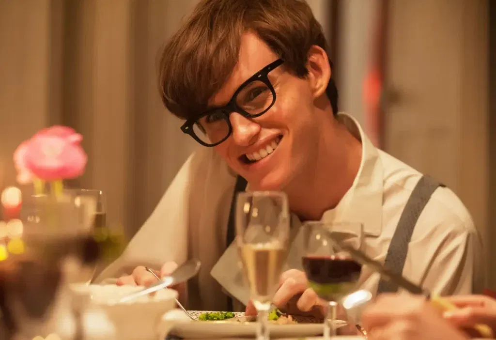 Close-up of a young man with tousled brown hair and thick black-framed glasses, grinning widely at the camera while seated at a candlelit dinner table. He wears a white shirt with grey suspenders and a light scarf, holding a spoon over a plate of food, with glasses of champagne and red wine and a small vase of pink roses in the foreground.