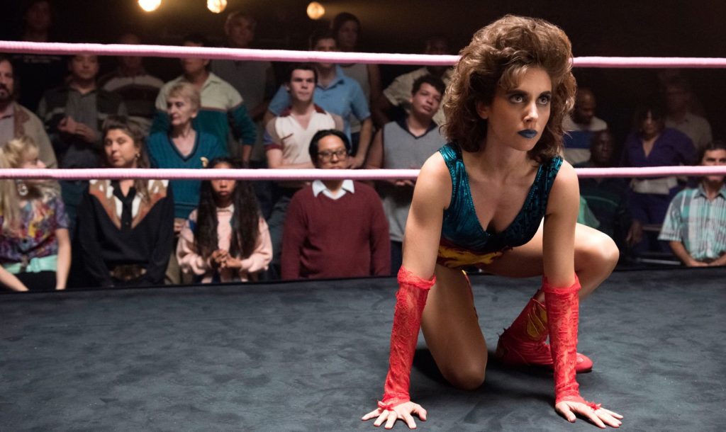  a woman with big, voluminous curly brown hair and blue lipstick crouches on all fours in a wrestling ring with pink ropes. She wears a teal metallic leotard with a yellow flame design, red lace arm warmers, and red boots, and stares intently off to the side. Behind the ropes, a crowd in 1980s-style clothing watches.