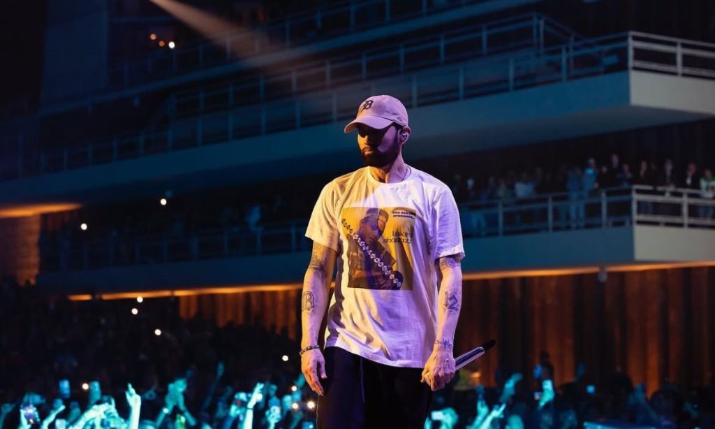 A bearded man in a light cap, a white tee with a yellow graphic print, and dark pants stands on a stage holding a microphone at his side, gazing off to the left with a serious look. Tattoos and a bracelet mark his arms. Behind him, a crowd fills the dim arena, many with raised hands and glowing phones, and tiered balconies rise above, crossed by a beam of stage light. He is Eminem