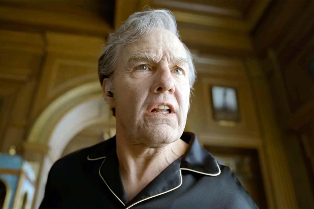 Low-angle close-up of an older man with swept-back grey hair and light eyes, scowling with his teeth bared in alarm or outrage. He wears black pajama-style loungewear with cream piping and a small earpiece. Behind him, a grand interior with wood-paneled walls, an arched doorway, and a framed picture glows in warm light. Tom Cruise in digger