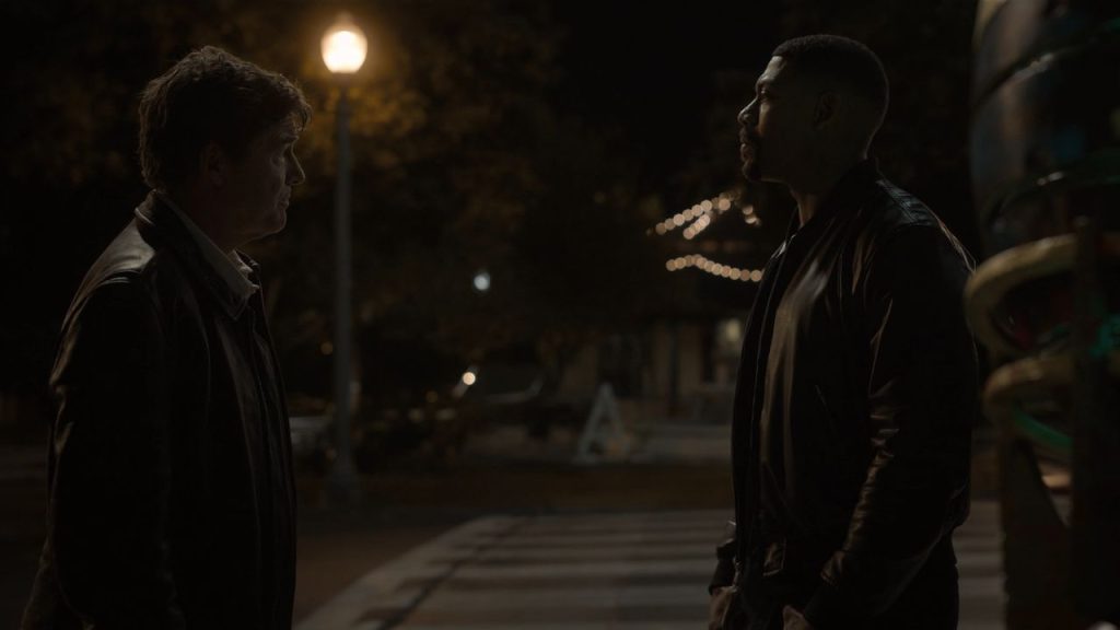 Two men face each other in profile on a dark park path at night, lit by a single glowing streetlamp. One wears a dark jacket over an open-collared shirt, the other a black leather bomber jacket.