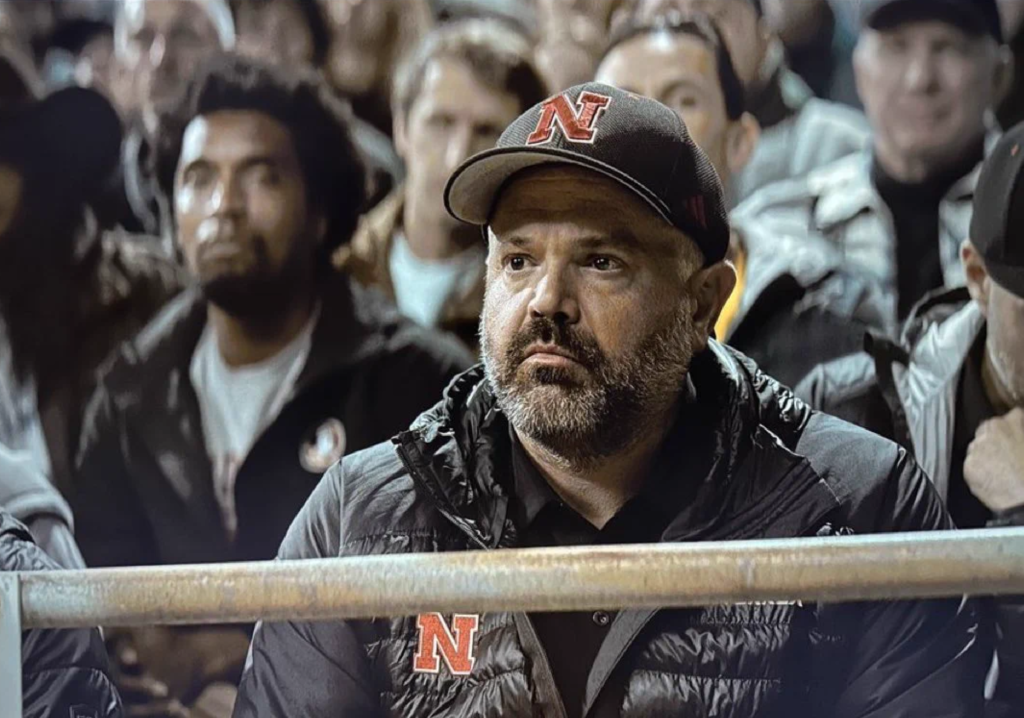 Lanterns Episode 8 Officially Turned Real-Life Football Coach Into DCU Canon 6 Close-up of a bearded man in a black baseball cap and black puffer jacket, both marked with a red block N logo. He sits behind a metal railing with a grim, watchful expression, and a blurred crowd of spectators fills the background. Matt Rhule cameo in lanterns episode 8