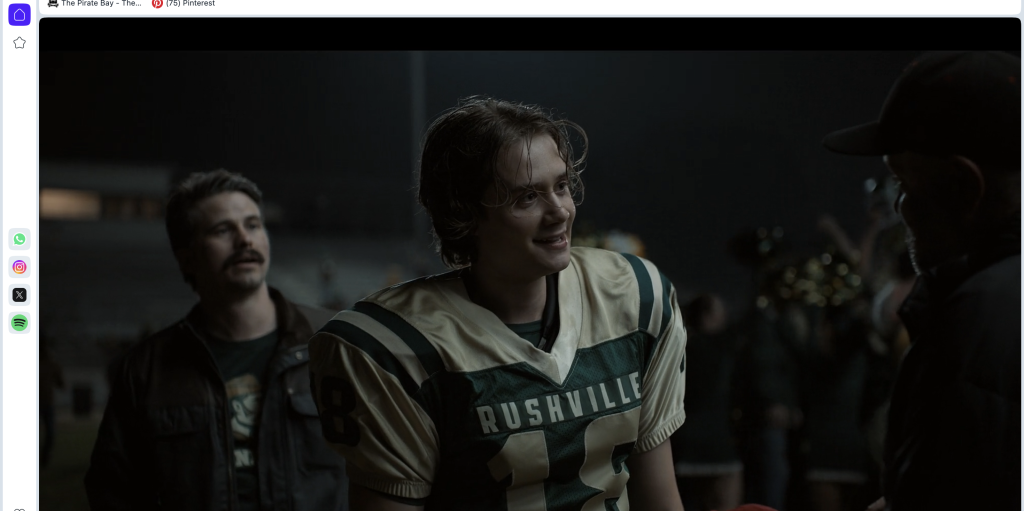 Lanterns Episode 8 Officially Turned Real-Life Football Coach Into DCU Canon 8 young man with tousled, damp hair and a faint smile wears a green and cream football jersey reading RUSHVILLE. A mustached man in a dark jacket and graphic tee stands behind him, and the shoulder of a person in a cap is in the right foreground. The stadium behind them is dark and blurred.