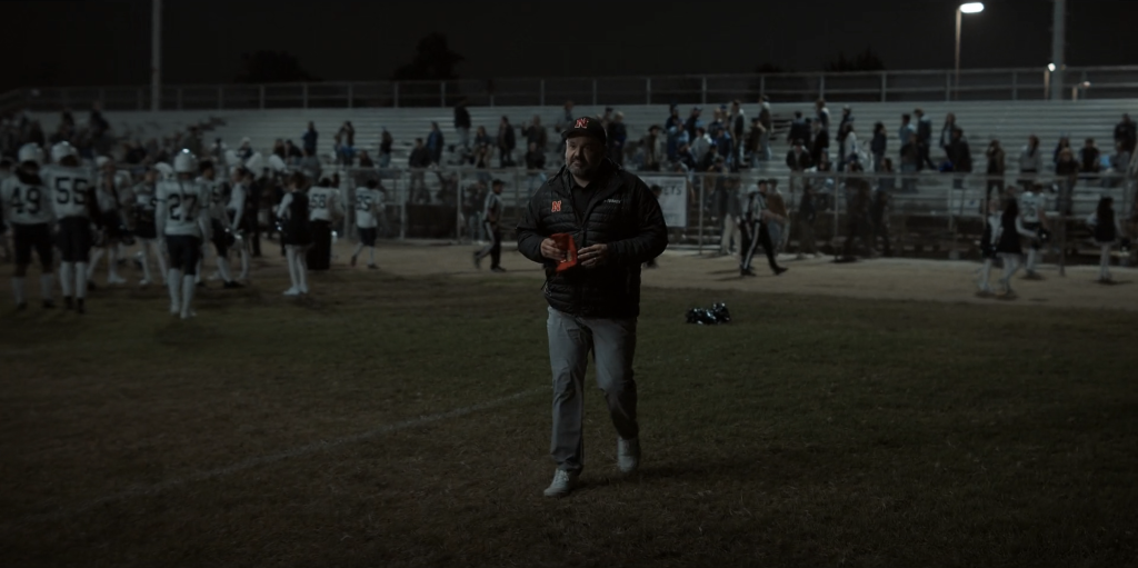 Lanterns Episode 8 Officially Turned Real-Life Football Coach Into DCU Canon 7 t night on a football field, a bearded man in a black cap and black puffer jacket with a red N logo walks toward the camera holding a small orange object. Behind him, players in white uniforms, officials and cheerleaders mill about in front of floodlit bleachers.