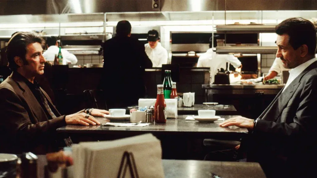 Two men sit facing each other across a table in a restaurant, in profile at opposite edges of the frame. On the left, a man with swept-back dark hair wears a brown jacket over a dark shirt, his hand resting on the table. On the right, a gray-haired man in a gray suit and white open-collar shirt, with a short graying beard, rests his hand on the table too. Two white cups, ketchup and condiments sit between them, and kitchen staff in whites work in the blurred, brightly lit kitchen behind. Martin Scorsese and Robert De Niro in heat
