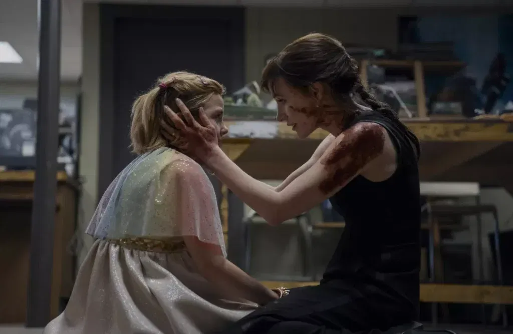 A woman in a black sleeveless dress kneels in a dim workshop-like room and cradles a young girl’s face in her hand. Dark red stains cover her hand, shoulder and cheek. She leans in close with a faint, tender smile. The girl, who has blonde hair in a small ponytail with pink clips, wears a shimmering rainbow cape over a pale sparkly skirt and gazes ahead. Wooden shelves and stools blur in the background.