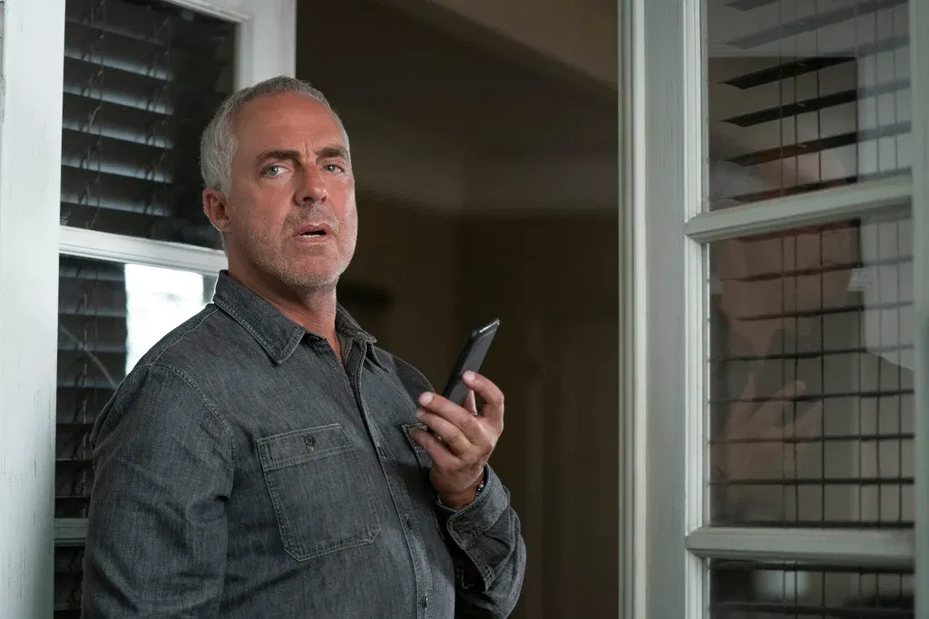 7 Best Prime Video Spinoffs, Ranked 6 A man with short gray hair and stubble stands in a doorway, holding a smartphone in one raised hand and glancing off to the side with a furrowed brow, his mouth slightly open as if mid-sentence. He wears a gray denim button-up shirt and a wristwatch. White-framed windows with slatted blinds surround him, and a dim hallway stretches out behind. Perhaps the best prime video spinoff ever