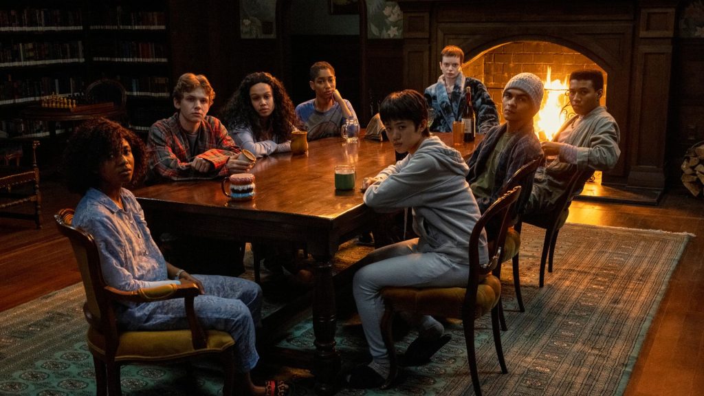 Eight teenagers in pajamas, robes and hoodies sit around a long wooden table in a dimly lit, book-lined library, all looking toward the camera with wary, solemn expressions. A fire burns in a brick fireplace behind them, and mugs, a glass jar and a wine bottle sit on the table. A chess set rests on a side table near the shelves, and a patterned green rug covers the floor. Mike Flanagan's show is better than carrie