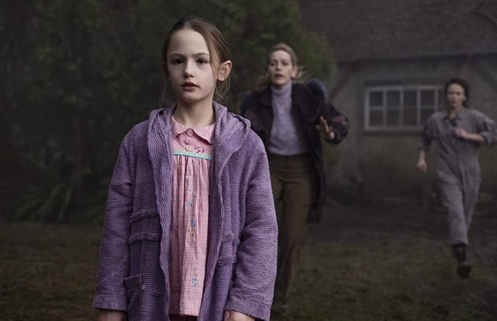 5 Mike Flanagan Horror Shows Better Than Carrie 2 A young girl in a purple chenille robe over a pink embroidered dress stands in the foreground of a misty, overgrown lawn, staring ahead with a blank, unsettling expression. Behind her, two women run toward her in alarm, one in a dark coat and lavender turtleneck with her hand outstretched, the other in a gray jumpsuit. A dim stone house with a paned window is barely visible through the fog.