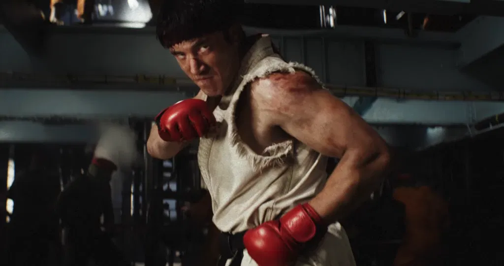 A cinematic still of Ryu from Street Fighter in a dim, industrial warehouse setting. He stands in a guarded fighting stance with his lead fist raised near his face and his rear fist low at his hip. He wears a frayed, sleeveless white gi, a black belt and red fingerless gloves. His shoulder is bruised and scuffed, and he stares intensely at something off-camera. Blurred figures are visible in the dark background.