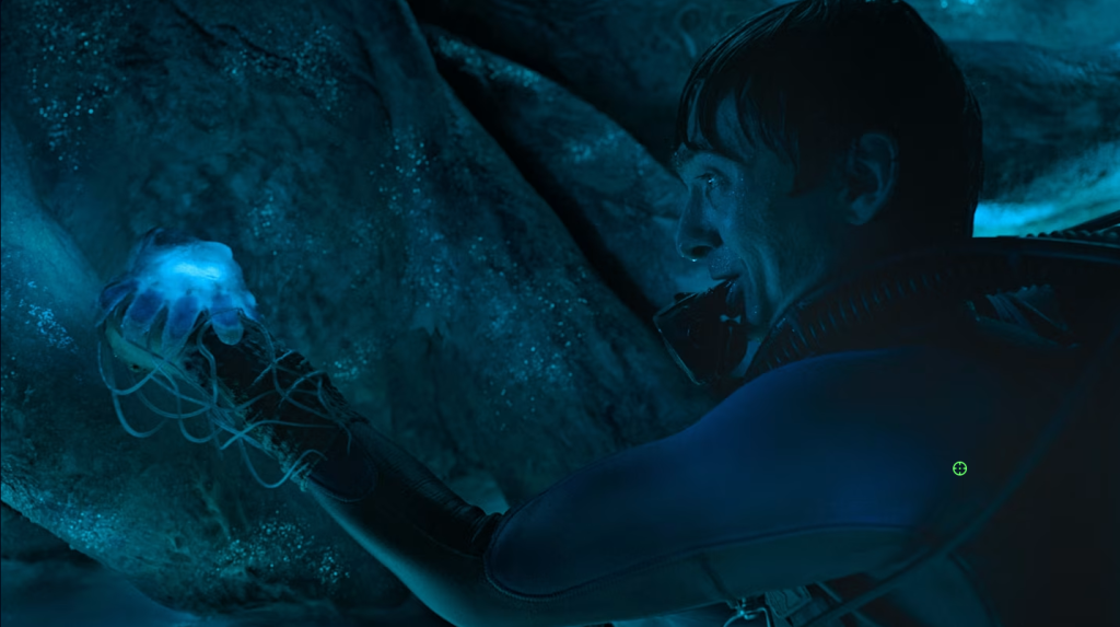 A diver in a blue and black wetsuit, seen in profile, stares at a glowing blue jellyfish held in his outstretched hand, its thin tendrils trailing down his forearm. His dark hair is wet and his regulator hangs loose near his mouth. Around him, dim blue-lit walls of rough, glistening flesh curve in, suggesting the inside of a whale.