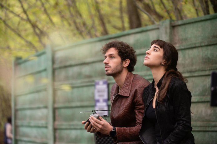 A man with dark curly hair and a short beard and a woman with long dark hair stand side by side, both looking up and into the distance. He wears a brown textured leather jacket and holds a small remote controller with an antenna. She wears a black leather jacket. Behind them are a weathered green concrete wall and leafy trees. A still from masterplan movie