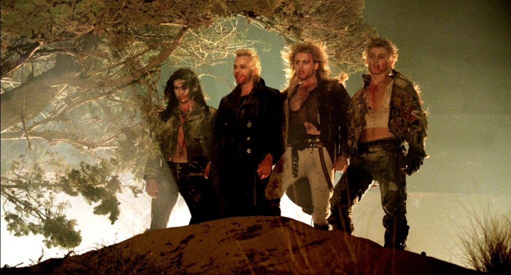  four young men in tattered leather and punk-style clothing stand shoulder to shoulder on a dirt mound at night, their faces and chests smeared with blood. The one on the left has long dark hair and a leopard-print collar, the second has spiky blond hair and a long black coat, the third has a big blond mane and a mesh top, and the one on the right has curly light hair and a patched, cropped jacket. Fog and a gnarled tree glow in pale green light behind them.