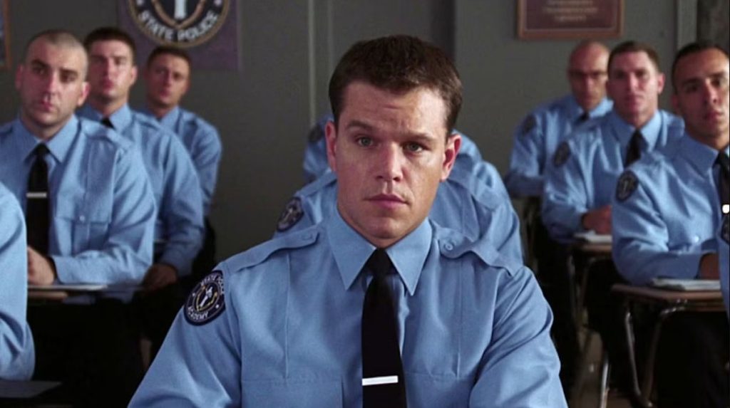 A young man with short brown hair sits at a classroom desk in a light blue uniform shirt with a black tie and a tie clip, staring ahead with a steady, serious expression. A State Police Academy patch is on his shoulder. Behind him, rows of other cadets in matching uniforms sit at desks, most looking forward, and a State Police banner hangs on the gray wall. Matt Damon in the departed