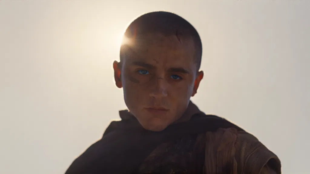  Part Three, with a closely shaved head, a scar on his forehead and blue-on-blue eyes, staring into the camera in silhouette as a bright sun flares behind his head.